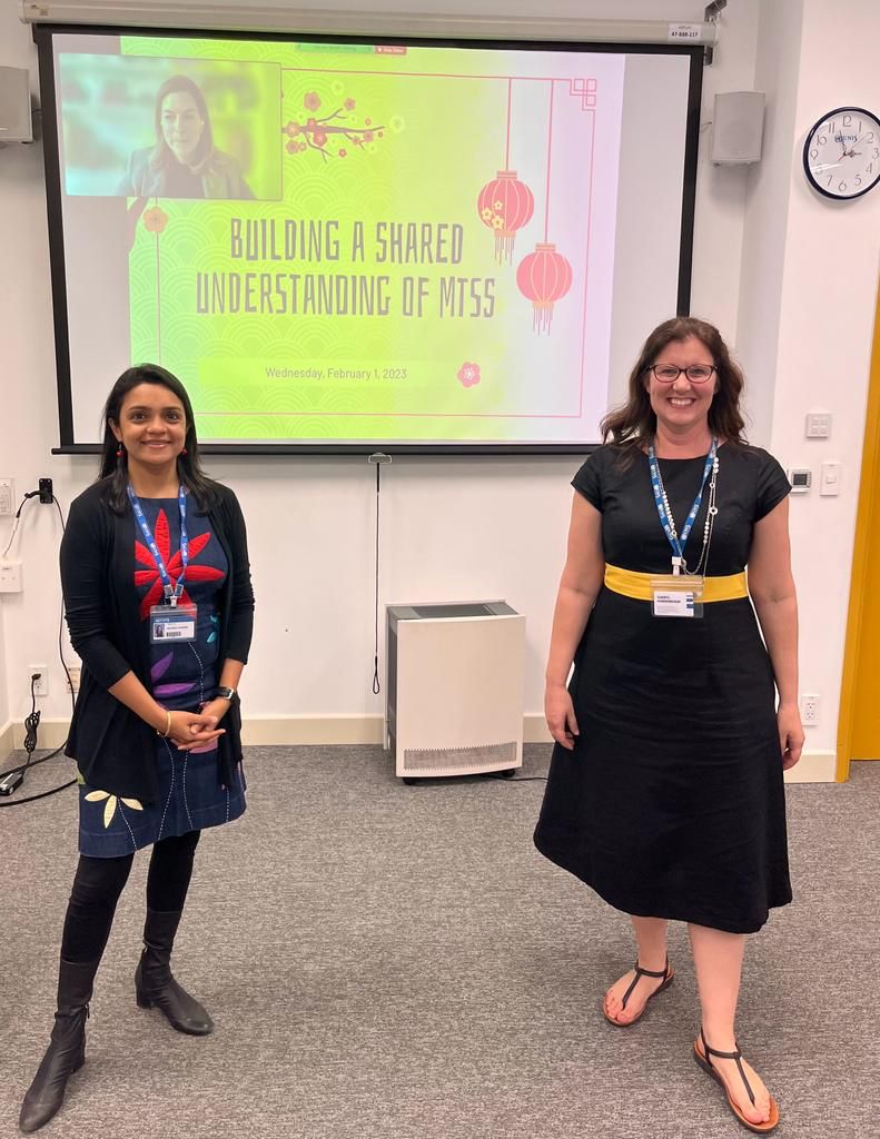 Two women stand in front of a screen with their co-presenter on screen and the title of their presentation: Building A Shared Understanding of MTSS.